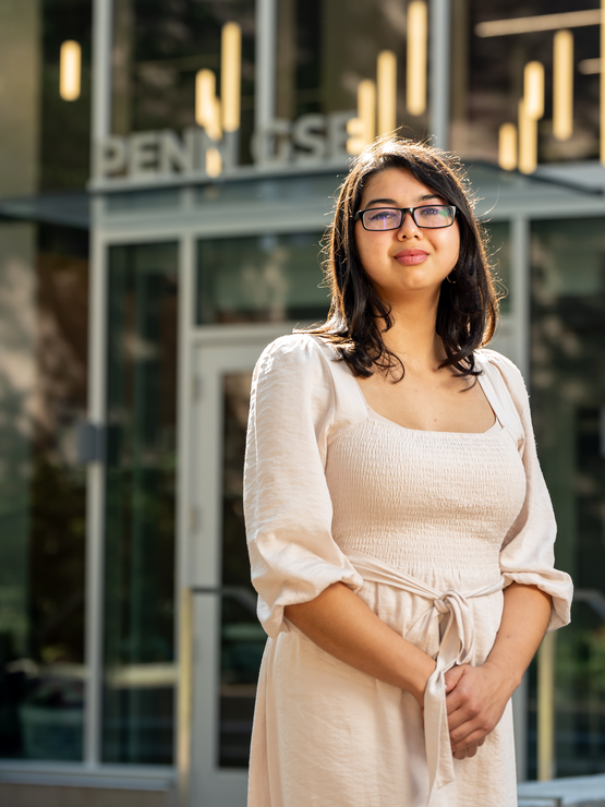 Ashley Zingillioglu in a cream-colored dress and glasses stands in front of the glass-walled entrance to Penn GSE