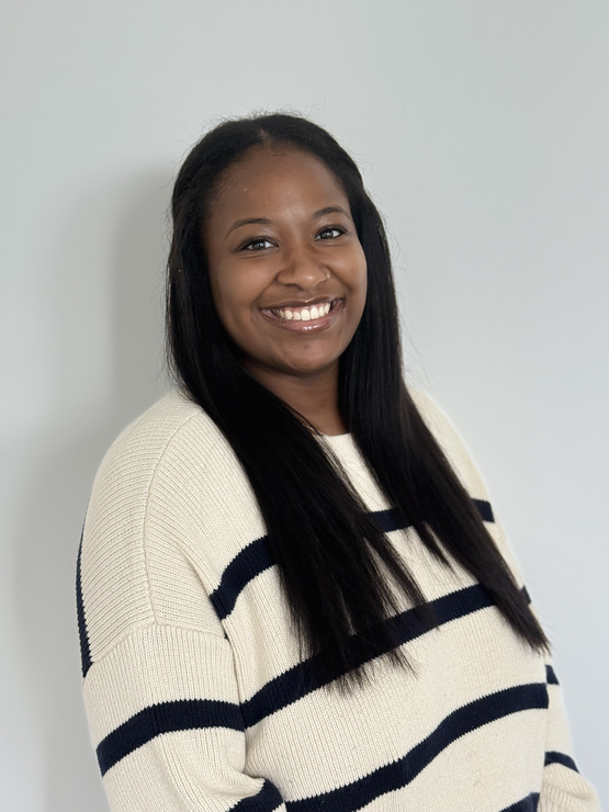 Rashelle smiles at the camera wearing a striped sweater and standing against a gray wall.
