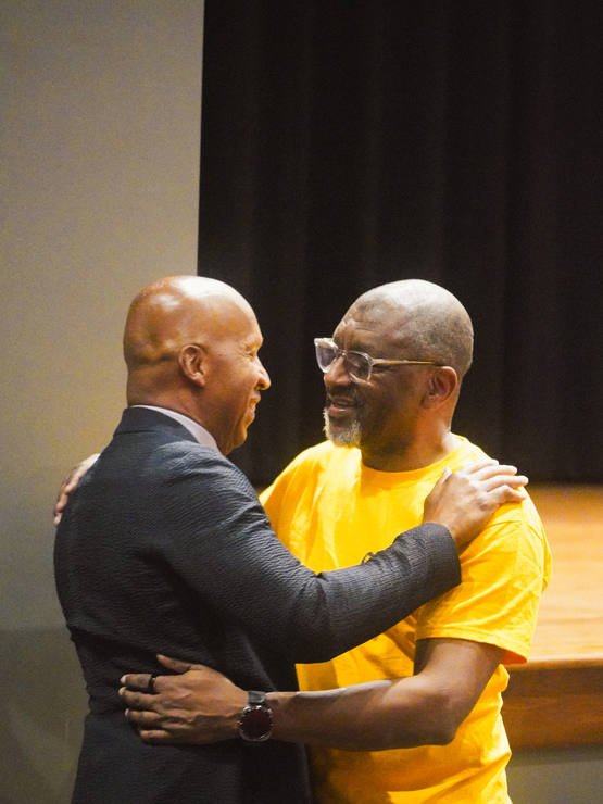 Bryan Stevenson, wearing a suit, hugs his brother Howard, wearing glasses and a bright yellow t-shirt. 