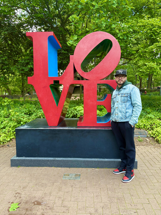Mark wears a jean jacket and Yankees cap standing next to the LOVE statue on Penn's campus.