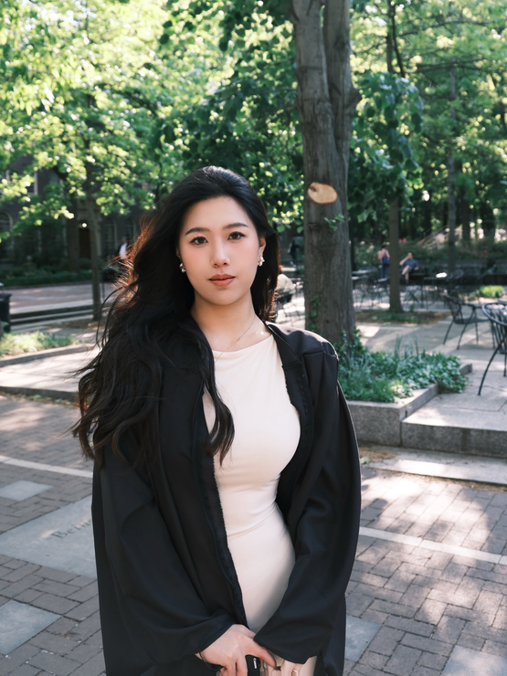 Xinyu wears her graduation gown over a white dress and poses in front of trees on the Penn campus