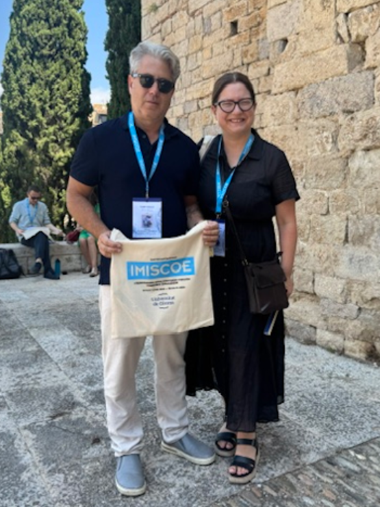Gerald and Maria stand together in front of an old bleached brick building in Spain wearing lanyards and holding a bag with the IMISCOE logo on it 