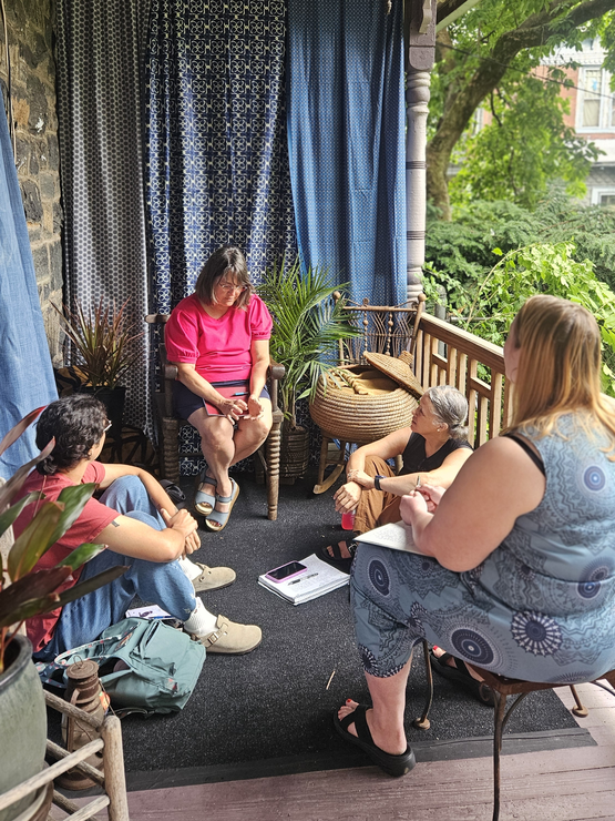 Someone sits on the ground and two other participants sit in chairs on a porch outdoors writing in notebooks.