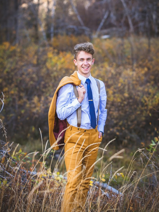 Zade wears a tie and mustard suit, holding the jacket over his shoulder as he stands in the woods in the fall. 