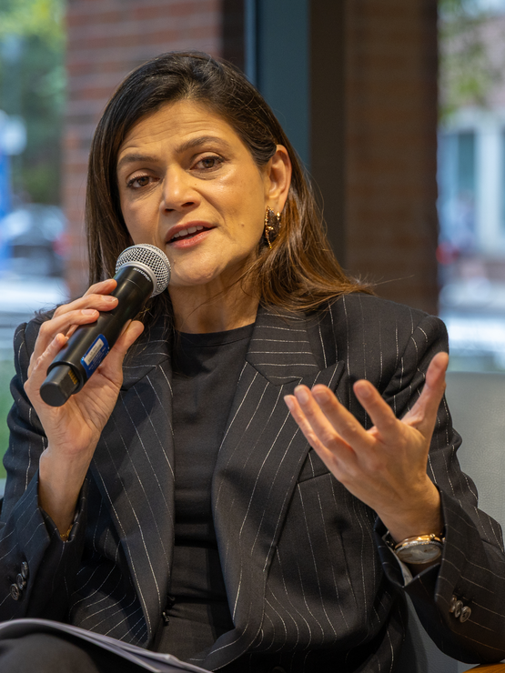 Mrs. Birla holds a microphone and gestures with her other hand while seated in front of a wall of windows.