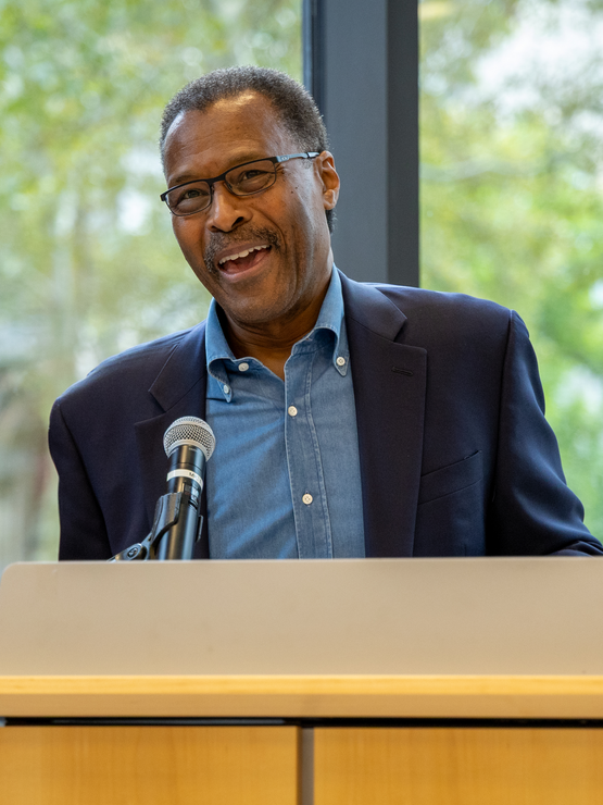 John Wilson smiling while speaking at a microphone on a podium.