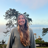 A woman smiles in front of backdrop of water, trees, mountaings, and blue skies with clouds