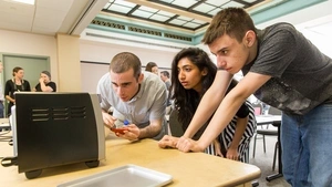 Three students gathered around a table, one pointing at a small appliance