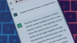 A smartphone is held in the center of the image with the ChatGPT website open on its browser. The phone is above a computer keyboard. You can see that the person has input “explain nuclear fusion in simple terms” as their prompt and the AI is generating an answer in return, illustrating how the program works for end-users.