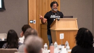 A man in a “We Need Black Teachers” t-shirt points to a screen while speaking at a podium in front of a crowd.