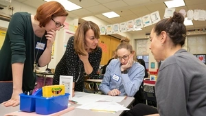 RMT trainers stand over a table and instruct two teachers, seated, in the teaching method