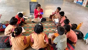 A teacher displays a chatbot on her phone screen while sitting cross-legged on a mat on the floor alongside a circle of her students during a Saturday Art Class in India.