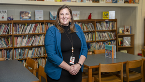 Julia Snyder stands in the middle of the Lea School library, smiling and wearing a blue cardigan. 