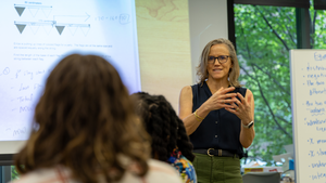 Caroline Ebby gestures with her hands in front of a screen showing math equations. The back of teachers heads, sitting in her class, are visible. 