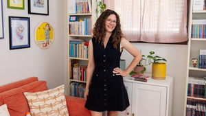 Katie Carella wears glasses and smiles with her hands on her hips inside her colorful office that is full of books