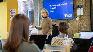 Julie Wollman stands in front of a class in front of a screen display that reads "The College nad University Presidency: Session 6." Students have their laptops open and are seated in two rows of desks. 