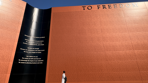 A person walking in profile appears small next to the grandeur of the monument, a large red wall that says "To Freedom" on the top 