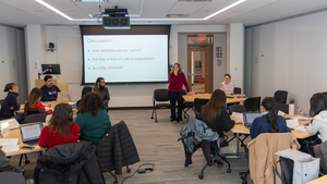 One teacher stands and another sits behind a table in front of a classroom of engaged students. Behind the teachers is a screen that reads: "Discussion: How restrictive are our norms? Is civility universal?"