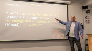 Seiji stands at a projection screen pointing at the words on it, which read "How can we bring the benefits of AI to people and places where technology and infrastructure are (almost) not available?"