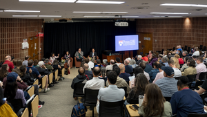 A full auditorium looks on as three professors are seated in chairs at the front of the room next to a screen with a Penn GSE logo on it. 