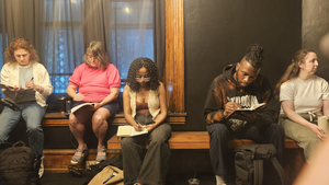 Five people sit along a bench in a windowsill indoors writing in journals
