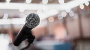 A view from behind a microphone with the conference room audience out of focus. 